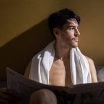 Shirtless man with towel reads newspaper in cozy bedroom setting, morning light.